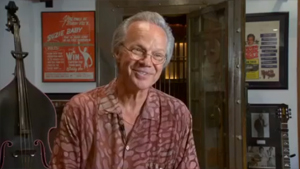 An older man smiling in front of a guitar.