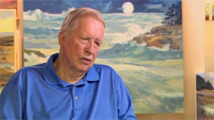 A man in a blue shirt sitting in front of paintings.