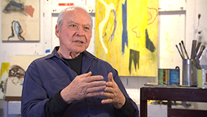 A man in a blue shirt sitting in front of a painting.