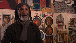 A man with dreadlocks standing in front of a room full of art.