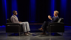 Two men in black chairs talking on stage.