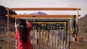 A woman is weaving on a wooden frame with mountains in the background.