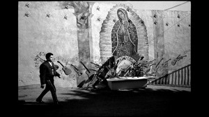 A black and white photo of a man walking past a mural of guadalupe.