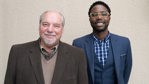 Two men standing next to each other in front of a wall.