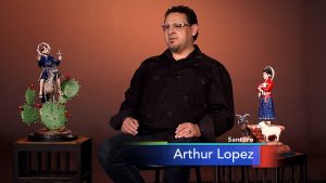 Arturo lopez, a mexican artist, sits on a stool next to two figurines.