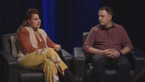 A man and a woman sitting in chairs on stage.