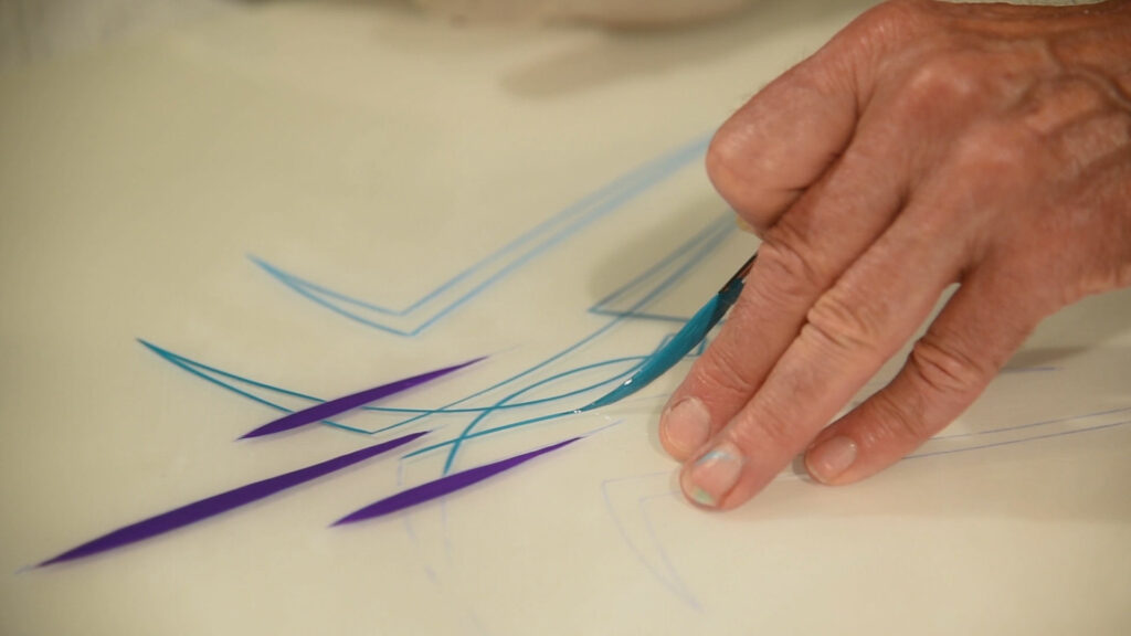 A hand holding a paint brush carefully paints blue and purple lines called "pinstriping"