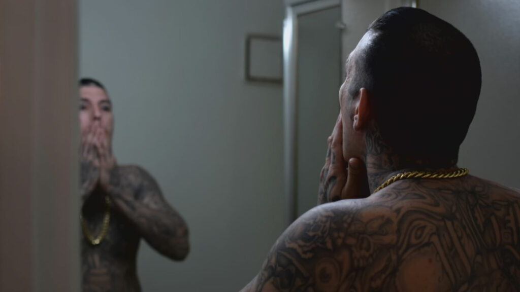 A person with tattoos holds their hands to their face while looking at their reflection in the mirror.