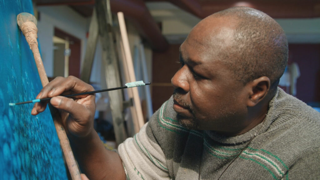 Talle Bamazi painting on a blue canvas on an easel.