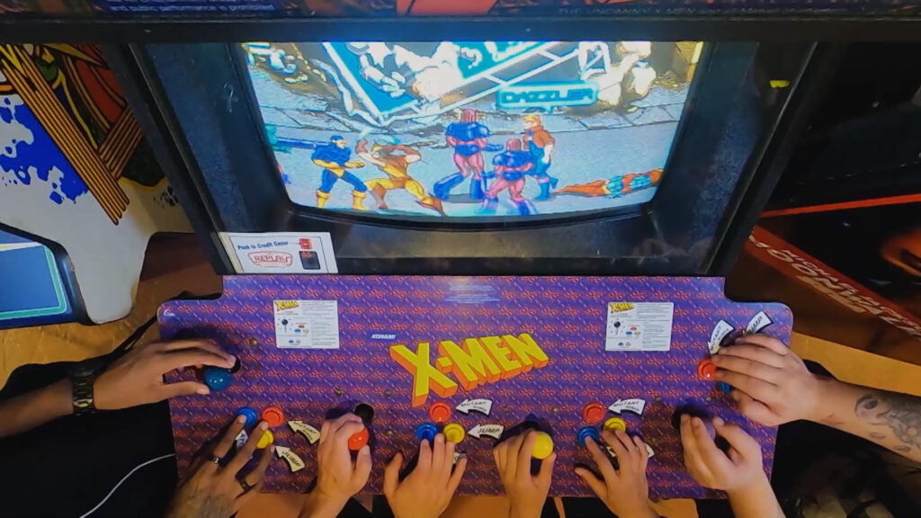 Multiple people gathered around an old arcade X-Men game. 