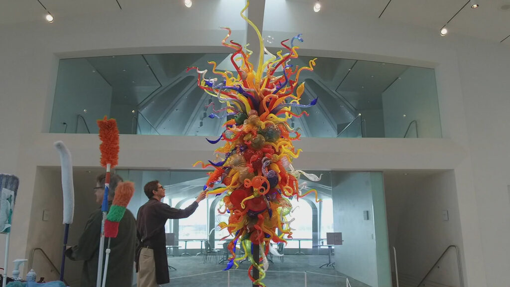 People dusting an elaborate glass art piece hanging from the ceiling.