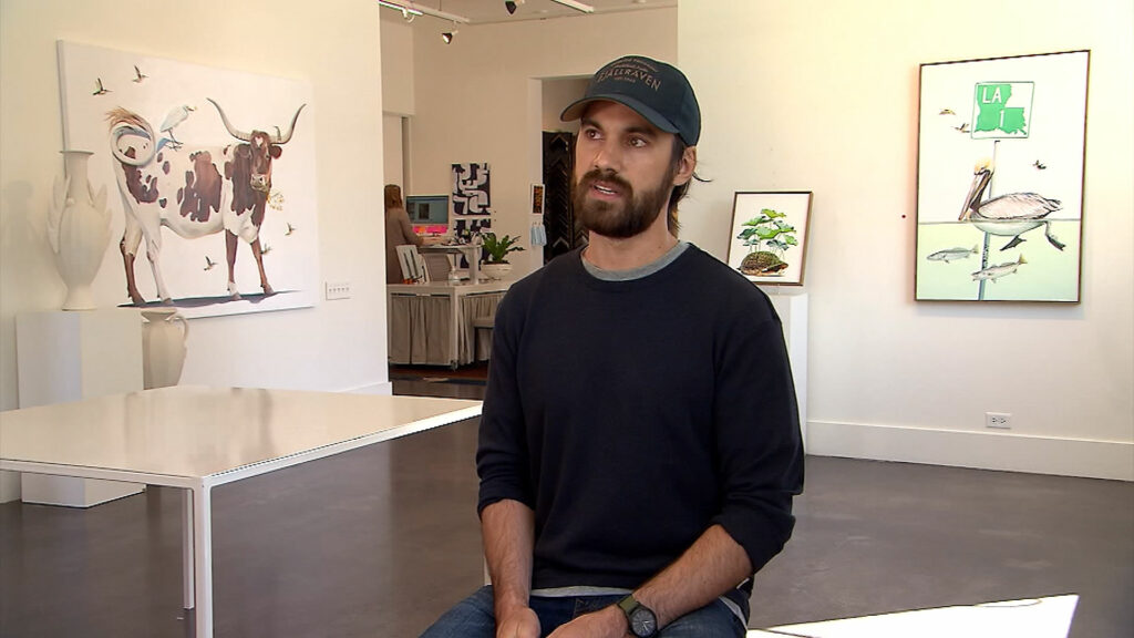 Chase Mullen sitting in a gallery surrounded by his work giving an interview.