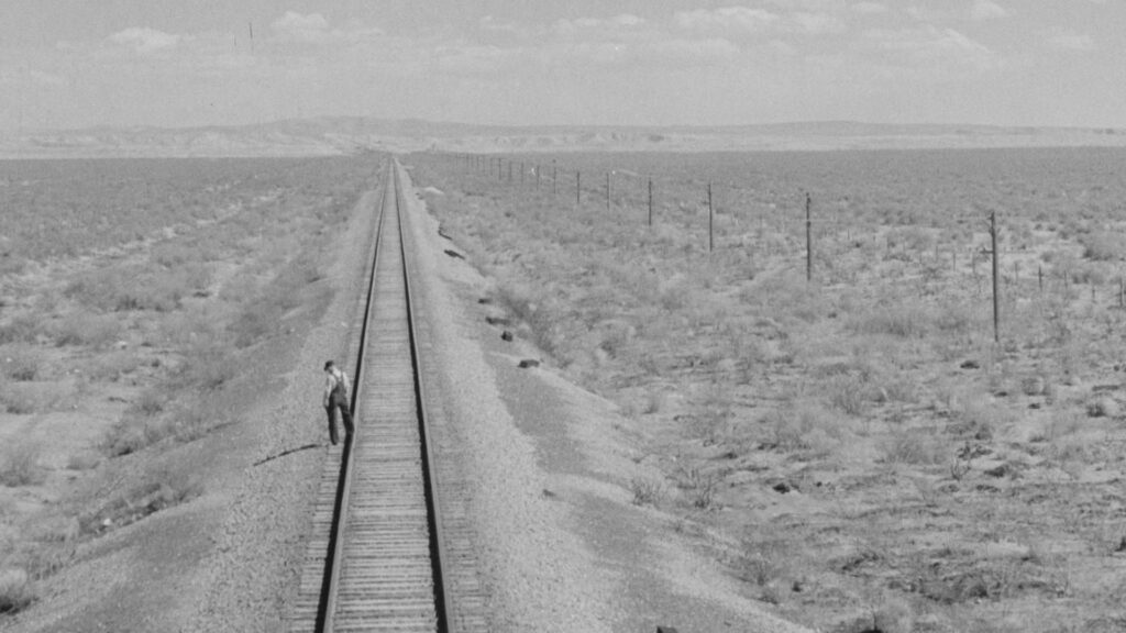 A black and white photo of a person walking across empty train tracks.