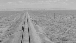 A black and white photo of a person walking across empty train tracks.