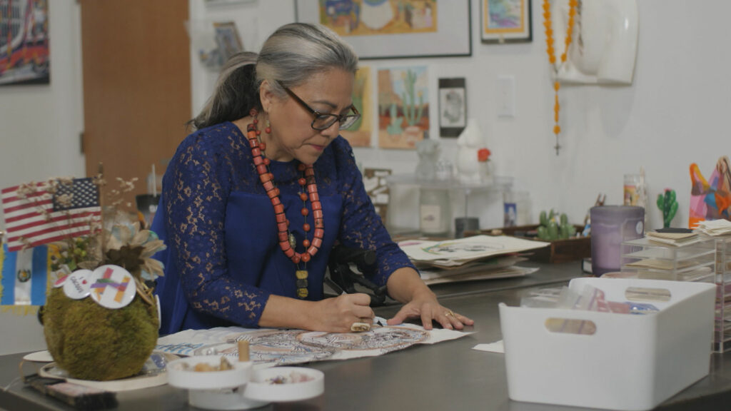 A woman sits at a desk and draws on a piece of paper.