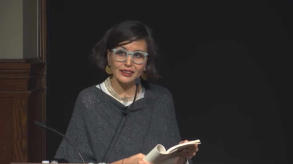 A woman in glasses reading a book in front of a microphone.