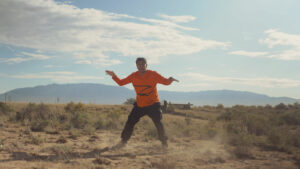 A man in an orange shirt flying a kite in the desert.
