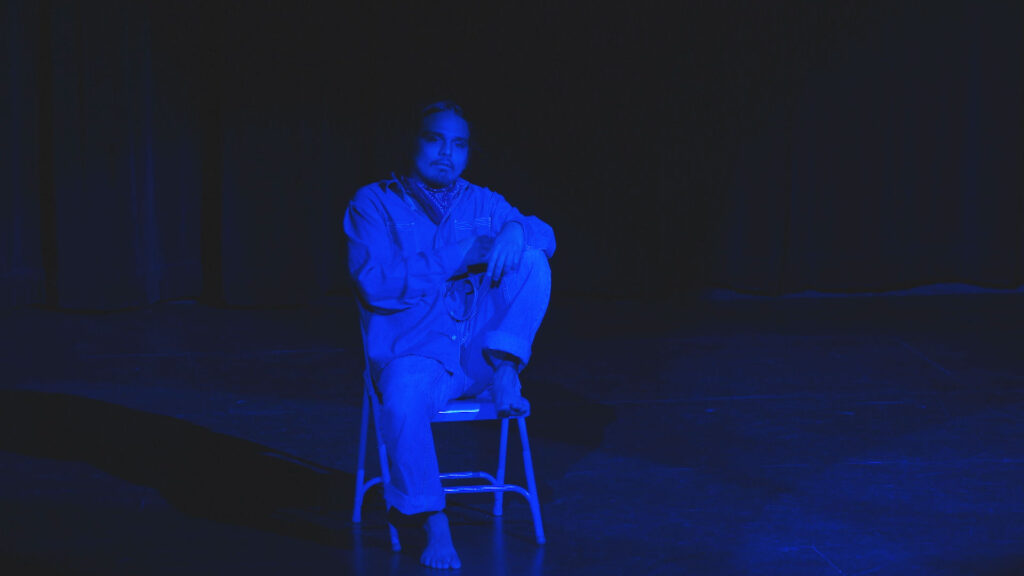 Person sitting on a chair under blue lighting on a dark stage.