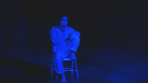 Person sitting on a chair under blue lighting on a dark stage.