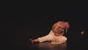 A performer in cultural attire engaged in a dramatic reading on a dark stage.