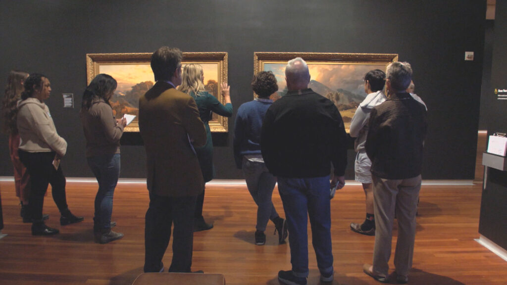 Visitors viewing landscape paintings in an art gallery room, some people are standing while others are seated on a bench.