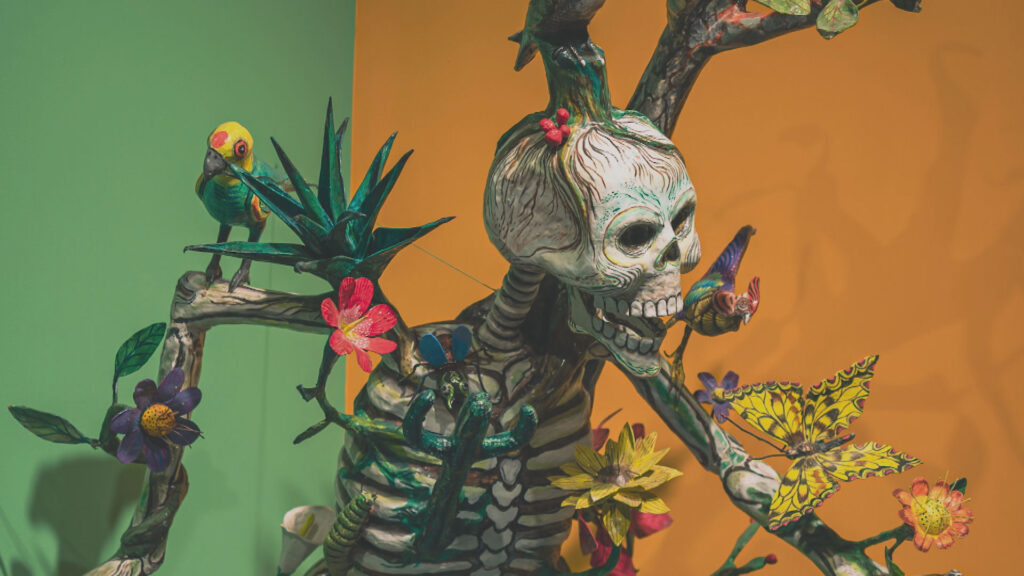 Skeletal figure adorned with colorful flora and fauna, including butterflies and a parrot, against a two-toned background.