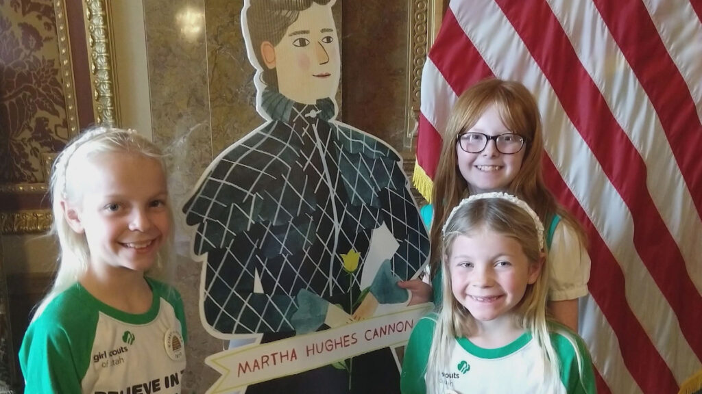 Three smiling children posing with a cardboard cutout of martha hughes cannon next to an american flag.