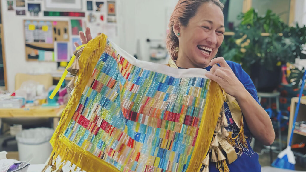A smiling woman displaying a colorful woven bag with fringe details.