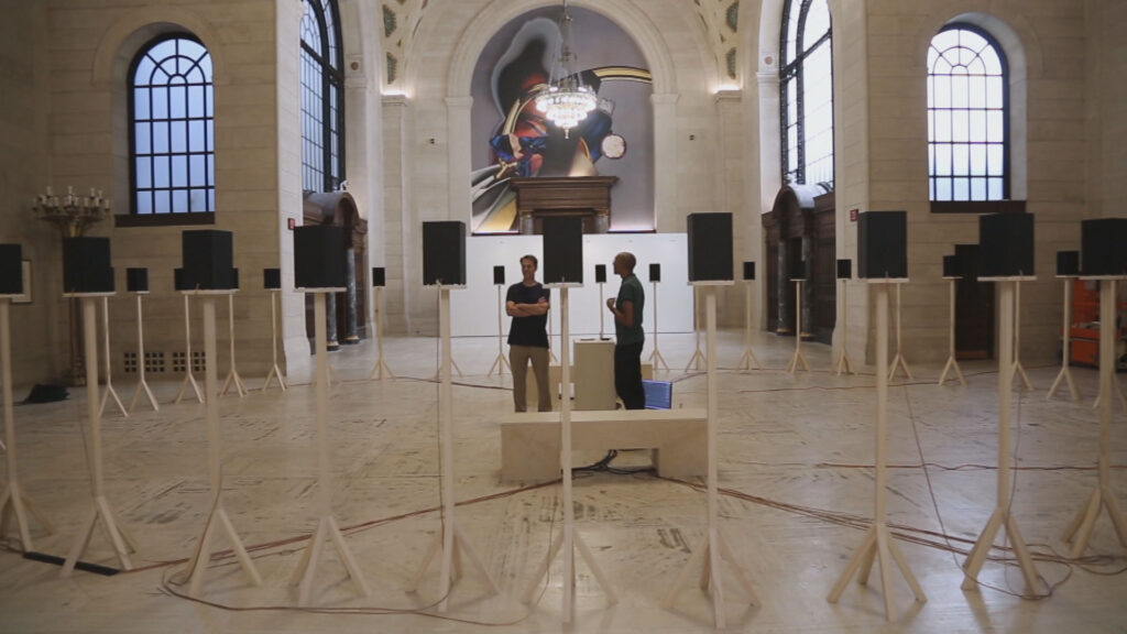Two individuals observe a sound installation featuring multiple speakers arranged in a historic interior space.