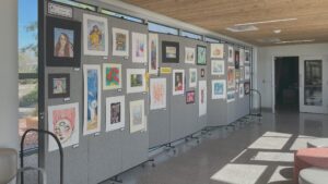 An indoor art exhibition with various paintings and drawings displayed on portable gray panels in a sunlit room with large windows.