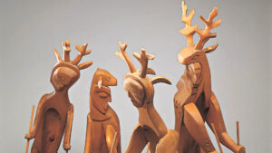 Four wooden sculptures featuring stylized human figures with deer antlers, standing on a neutral background.