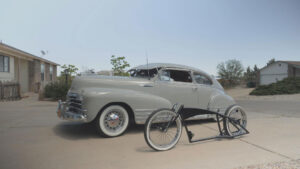 A vintage gray car is parked in a driveway next to an incomplete bike frame with wheels.