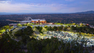 A large illuminated parking lot fills with cars outside a distant building at dusk, set against a backdrop of rolling hills and a twilight sky.