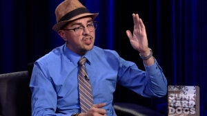 A man wearing a fedora, glasses, and a blue shirt with a striped tie is gesturing with his right hand while speaking. A "Junk Yard Dogs" logo is visible in the lower right corner.