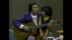 An adult in a purple jacket is sitting with a child, playing an acoustic guitar, while both look in the same direction.