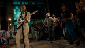 Musician plays electric guitar on stage while audience dances and claps in a dimly lit indoor venue.