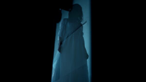 Silhouette of a person in a long dress standing in a narrow beam of light against a dark background.