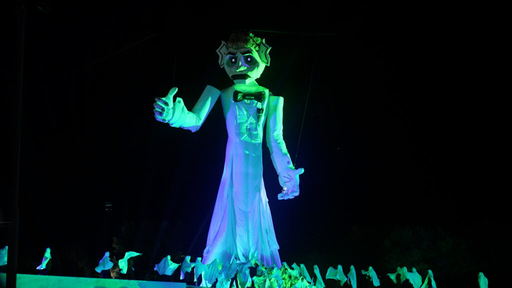A large, glowing puppet figure in a white robe with outstretched arms is illuminated against a dark background. Smaller figures with wings are at its base.