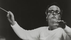 A person wearing glasses conducts with a baton, dressed in a white long-sleeve shirt against a dark background.