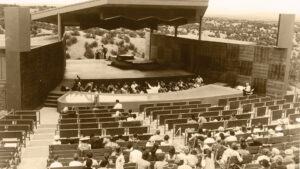 Outdoor amphitheater with audience watching an orchestra perform on stage, surrounded by a scenic landscape.
