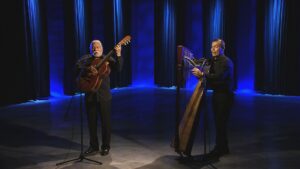 Two musicians perform on stage, one playing a guitar and the other a harp, under blue lighting.