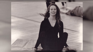 A woman sitting on a mat, dressed in a dark outfit, with hair flowing back, inside a large indoor space.