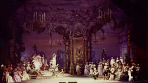 Ornate stage set with actors in period costumes, featuring chandeliers and detailed architecture, depicting a historical or fantasy scene.