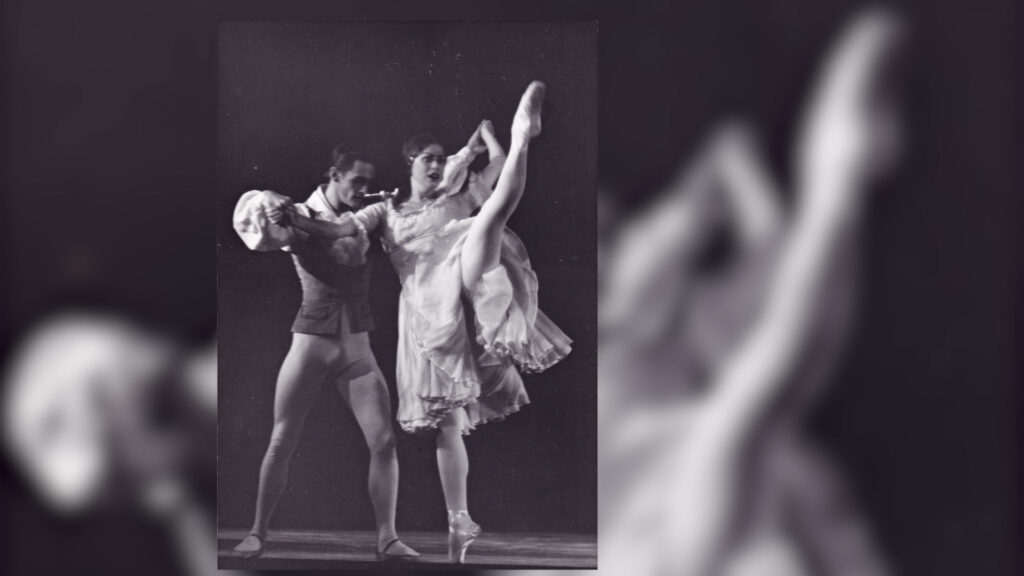 Two ballet dancers perform on stage; the female dancer is mid-leap with her leg raised high, while the male dancer supports her. They are dressed in classical ballet costumes.