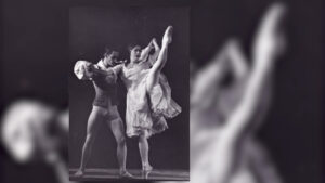 Two ballet dancers perform on stage; the female dancer is mid-leap with her leg raised high, while the male dancer supports her. They are dressed in classical ballet costumes.