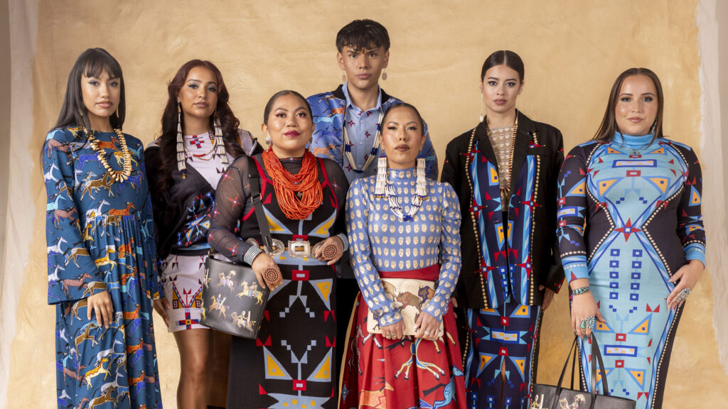 Seven people in colorful, patterned outfits stand together against a beige background.