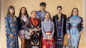 Seven people in colorful, patterned outfits stand together against a beige background.