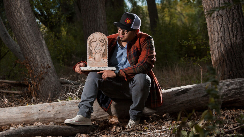 Person sitting on a fallen tree in a forest, holding a carved wooden piece depicting a face.