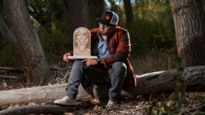 Person sitting on a fallen tree in a forest, holding a carved wooden piece depicting a face.