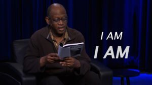 A person in glasses reads from a book while seated against a dark curtain. The words "I AM I AM" are displayed beside them.